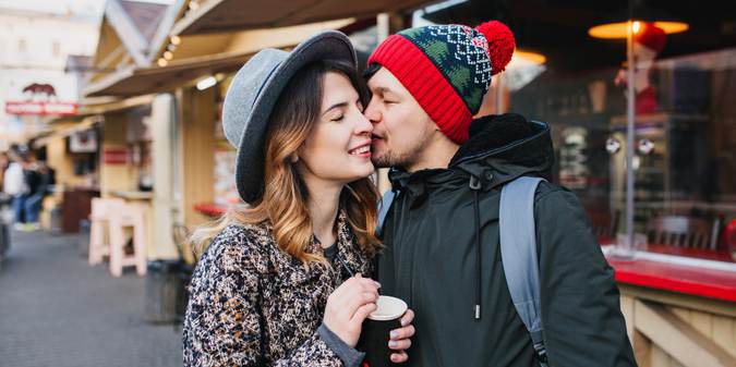 Couple sharing a warm kiss at an outdoor market, holding coffee, wearing winter outfits in a cozy street setting